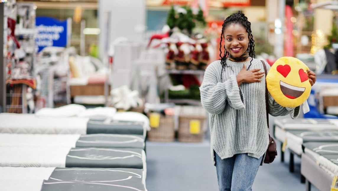 A picture of a woman holding a smiley face pillow while standing by mattresses.