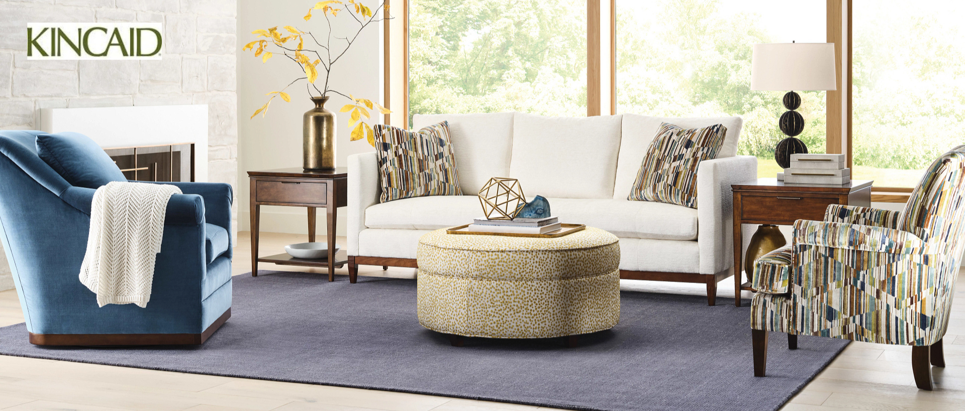 Living room with white sofa, blue arm chair and multi colored arm chair with a round yellow ottoman in the center