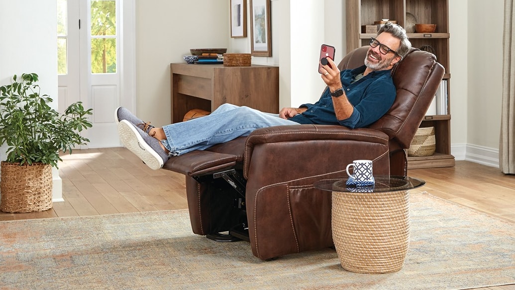 Man relaxing in a brown leather recliner looking at this phone and smiling